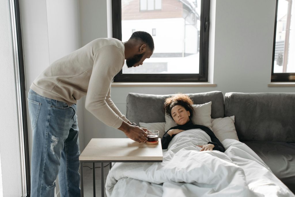 A man attentively cares for his sick partner by bringing her tea as she rests on the sofa.